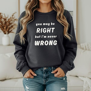May include: A dark charcoal gray crewneck sweatshirt featuring white text that reads you may be RIGHT but i'm never WRONG. The long-sleeved pullover has a ribbed neckline, cuffs, and hem, providing a standard fit for casual wear.