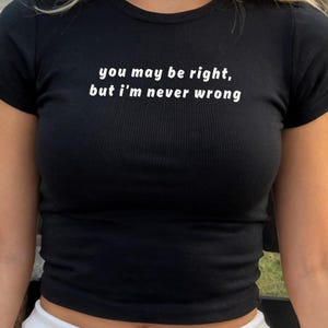 May include: A black short-sleeved ribbed t-shirt featuring the white printed text "you may be right, but i'm never wrong" centered on the chest. The garment has a crew neckline and a fitted silhouette with a straight hemline at the waist.