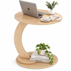 May include: A light wood C-shaped side table with a circular top and base. The table features a curved support column connecting the two levels. The top surface holds a laptop, a white vase with greenery, a coffee cup, and a notebook with a pen.