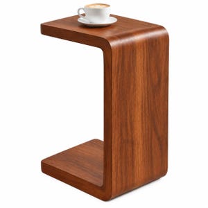 May include: A C-shaped side table crafted from dark brown wood with a smooth, continuous curved design. A white ceramic cup filled with coffee sits on the flat top surface of the table, which features a visible wood grain pattern throughout.