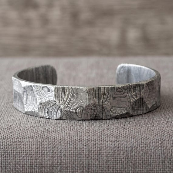Hand-forged Damascus steel cuff with the hypnotic layered-metal grain pattern — this is actual blacksmithing craft, zero chance you've seen this in a store.