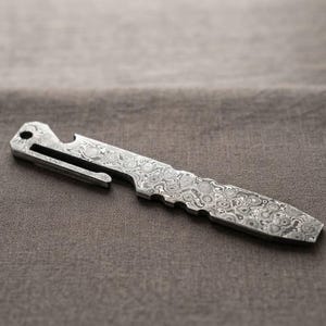 May include: A multi-functional pocket tool crafted from Damascus steel with a distinct swirling grey and silver pattern. The tool features a flat, pointed end, a series of notches along one edge, a bottle opener cutout, and a lanyard hole.