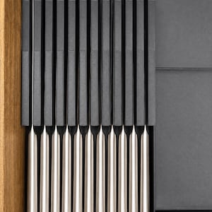 May include: A set of twelve knives with matte black handles and silver-toned metal blades arranged vertically in a dark gray drawer organizer. The knives feature a uniform design with rounded handle ends and straight, slender blades aligned side by side.