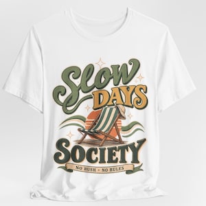 May include: A white t-shirt featuring a graphic print with the text "SLOW DAYS SOCIETY" and "NO RUSH • NO RULES". The design includes a green and white striped beach chair, a sun, and small star accents in shades of green, orange, and tan.