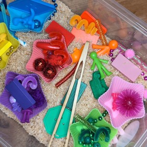 May include: A clear plastic sensory bin filled with light brown rice and various colorful items including star-shaped silicone cups in yellow, red, purple, pink, and green. The bin contains small plastic figures, pom-poms, beads, and wooden tongs.