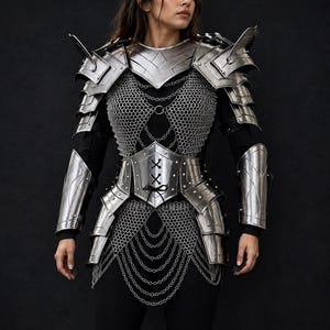 May include: A silver metal armour set featuring layered shoulder pauldrons, a chest plate with integrated chainmail, a waist corset with black lace-up detailing, and matching forearm bracers. The armour includes decorative hanging silver chains.