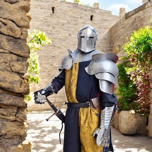 May include: A suit of armor featuring a polished steel helmet, shoulder pauldrons, and articulated gauntlets. The ensemble includes a black and gold patterned tunic with a chainmail coif, a black belt, and a sword held in a black scabbard.