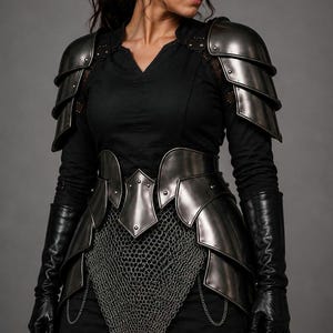 May include: A set of dark metallic plate armour featuring layered shoulder pauldrons, a waist-cinching cuirass with pointed accents, and a chainmail skirt. The ensemble is worn over a black long-sleeved shirt and includes long black leather gloves.