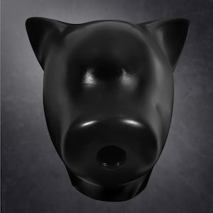 May include: A matte black, stylized pig head sculpture featuring pointed ears and a prominent snout. The snout contains a circular opening with visible internal threading, creating a smooth, solid-colored form with a distinct animal silhouette.