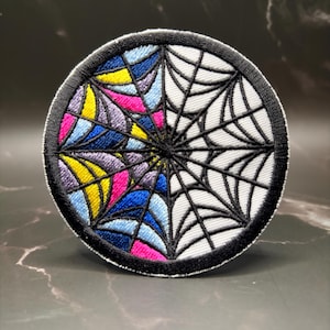 May include: A circular embroidered patch featuring a spider web design. The left half displays vibrant segments of yellow, pink, light blue, and dark blue, while the right half features a classic black and white spider web pattern.