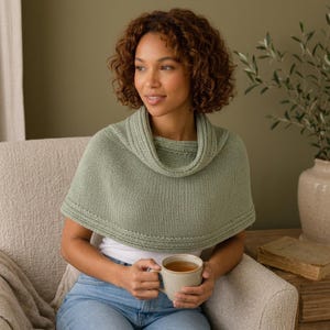May include: A sage green knitted cowl neck poncho featuring a ribbed texture and a wide folded collar. The garment is worn over a plain white top and blue denim jeans, showcasing a simple knit construction with a rounded hemline and detailing.