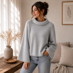 May include: A light gray knit sweater featuring a wide, folded turtleneck collar and long, loose bell sleeves. The garment has a soft, textured appearance with ribbed detailing at the cuffs and hem, paired with light blue denim jeans.