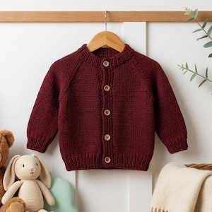 May include: A deep burgundy long-sleeve knit cardigan featuring a round neckline and five light brown wooden buttons down the centre front. The garment has ribbed cuffs and a ribbed hem, showcasing a consistent textured stitch pattern throughout.