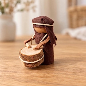 May include: A peg doll figure draped in brown fabric with a matching head covering, featuring a natural wood head. The figure holds two small wooden sticks and is positioned behind a drum made from a circular slice of wood with bark edges.