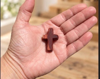 May include: A small, smooth, reddish-brown wooden cross with rounded edges. The cross is shaped with a vertical bar longer than the horizontal bar, featuring a consistent dark wood grain texture throughout its surface, held in an open palm.