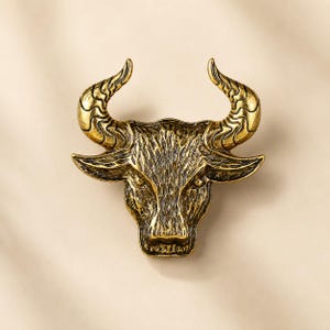 May include: A metallic gold-toned bull head ornament featuring a textured finish on the face and intricate swirling patterns carved into the curved horns. The design captures the detailed facial features of a bull with a prominent snout and ears.
