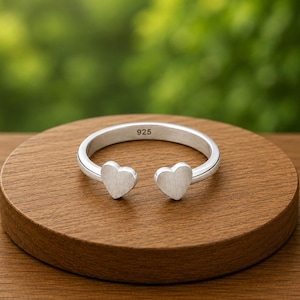 May include: A silver open-style ring featuring two small heart-shaped ends. The inner band of the ring is clearly stamped with the number 925, indicating sterling silver composition. The ring has a smooth, matte finish and a simple, clean design.