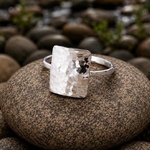 Könnte beinhalten: Ein Silberring mit einer großen, rechteckigen, gehämmerten Metalloberfläche, die eine texturierte und reflektierende Optik aufweist. Die Ringschiene ist schmal und zeigt ebenfalls ein passendes gehämmertes Finish für ein stimmiges Design.