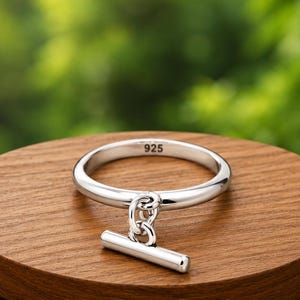 May include: A polished silver ring featuring a smooth, thin band with a small, dangling bar charm attached by a circular link. The interior of the band is clearly stamped with the hallmark 925, indicating sterling silver composition.