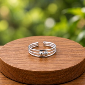 Può includere: Un anello color argento caratterizzato da tre fasce parallele aperte sul retro. La parte anteriore dell'anello è decorata con due piccole perle sferiche lisce, posizionate fianco a fianco e centrate sulle tre sottili fasce metalliche.