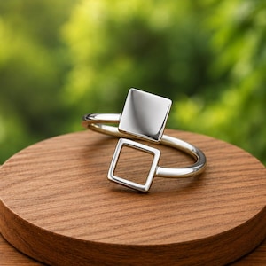 Puede incluir: Un anillo de plata con dos elementos cuadrados, uno sólido y otro abierto, unidos a una banda fina y pulida. El diseño presenta una disposición geométrica con un acabado metálico reflectante, creando una apariencia limpia y equilibrada.