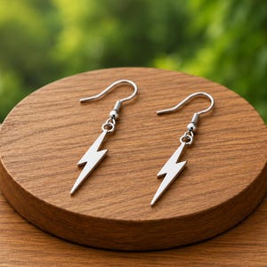 Könnte beinhalten: Ein Paar Ohrringe aus silberfarbenem Metall mit einem Blitz-Anhänger, der an einem Standard-Ohrhaken befestigt ist. Die Ohrringe haben eine glatte, polierte Oberfläche und ein schlichtes, klares Design, das vertikal am Ohrhaken hängt.