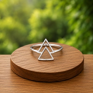 Pode incluir: Um anel de tom prateado com um design geométrico composto por dois triângulos abertos sobrepostos. A banda é fina e circular, suportando o ponto focal triangular que se assenta verticalmente no topo da circunferência do anel.