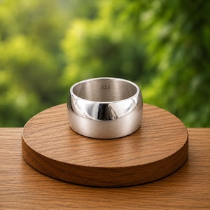 May include: A wide, polished silver band ring with a smooth, reflective surface. The interior of the ring is clearly stamped with the hallmark 925, indicating sterling silver composition. The ring features a simple, unadorned, and cylindrical design.