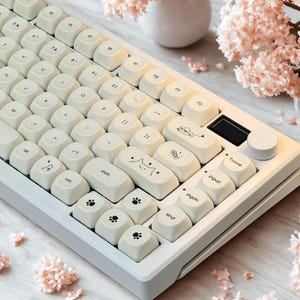 Puede incluir: Un teclado mecánico blanco con teclas redondeadas y caracteres impresos en negro, incluyendo iconos de gatos como huellas, una cara de gato y un pez. El teclado cuenta con una perilla de volumen circular y una pequeña pantalla rectangular.