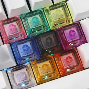 Könnte beinhalten: Eine Sammlung von neun quadratischen, durchscheinenden, mehrfarbigen mechanischen Tastaturkappen, die in einem Raster angeordnet sind. Die Tastenkappen haben eine klare, glänzende Oberfläche in den Farben Pink, Türkis, Gelb, Blau, Schwarz, Lila, Transparent, Orange und Rot.