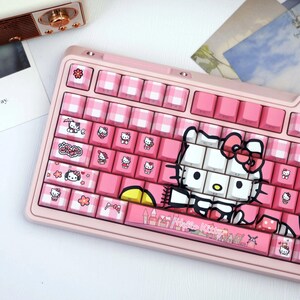 May include: A pink mechanical keyboard featuring custom keycaps with a pink and white gingham pattern. Several keys display Hello Kitty character graphics, and the spacebar features a printed castle scene with the text Hello Kitty in pink.