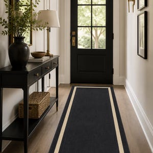 May include: A long rectangular runner rug featuring a solid black centre bordered by a thick cream-coloured frame. The rug is positioned on a light wood floor in a hallway, leading towards a black door with glass panes and brass hardware.
