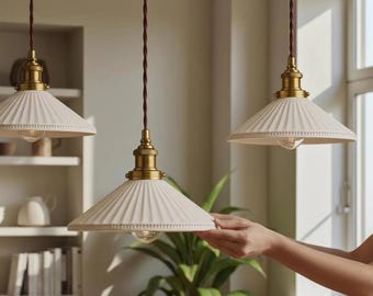 May include: Three pendant lights featuring white, fluted, conical ceramic shades with brass-toned hardware and brown twisted fabric cords. Each light fixture has a textured rim along the edge of the shade and a clear bulb visible underneath.