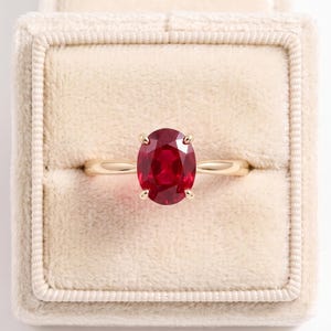 Puede incluir: Un anillo de oro con una gema de rubí rojo intenso de corte ovalado sujeta por cuatro garras. El anillo está centrado dentro de un joyero cuadrado forrado de terciopelo beige que tiene una superficie suave y texturizada con la tapa detrás.