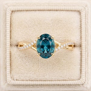 May include: A gold ring featuring a central oval-cut teal gemstone held by four prongs. The band is split into two intertwining strands, each accented with a row of small, round, clear pavé-set diamonds that extend from the center stone outward.