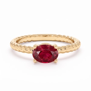Op de afbeelding: Een gouden ring met een centrale ovaal geslepen dieprode edelsteen, vastgehouden door vier pootjes. De ringband heeft een opvallende gedraaide touwstructuur, wat een doorlopend patroon creëert. Het metaal heeft een gepolijste, glanzende afwerking.