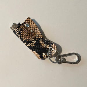 Puede incluir: Una funda para encendedor con un patrón de piel de serpiente en tonos beige y negro, acentuada con una fila de tachuelas metálicas plateadas en el lateral. La funda incluye una anilla metálica plateada y un mosquetón a juego.