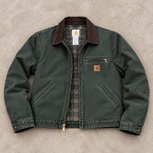 May include: A dark green canvas jacket featuring a brown corduroy collar, a full-length front zip, and a plaid interior lining. The jacket includes a zipped chest pocket with a Carhartt logo patch and two lower front pockets.