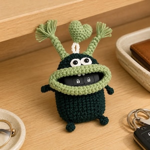 May include: A dark green crochet key fob cover shaped like a creature with a wide light green mouth, two antennae topped with tassels, and a small light green heart attached to the top. The cover holds a black car key fob with lock icons.