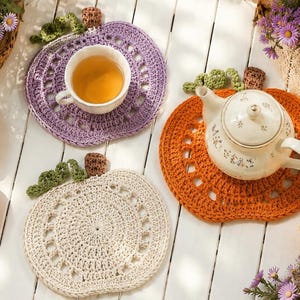 May include: Three round crochet mats shaped like pumpkins in lavender, orange, and cream. Each mat features a green crocheted vine and a brown stem. The lavender mat holds a white teacup, and the orange mat holds a white floral teapot.