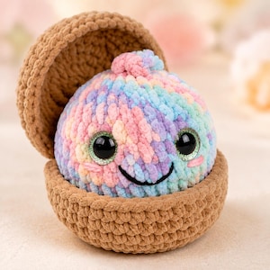 Puede incluir: Un juguete de peluche de ganchillo redondo y multicolor con hilo rosa, azul y morado pastel, con grandes ojos de seguridad negros y una sonrisa bordada. El peluche se asienta dentro de una concha de ganchillo marrón texturizada de dos piezas.