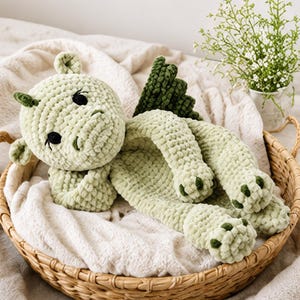 May include: A light green crocheted dragon plush toy with dark green wings and dark green accents on its paws. The creature has black safety eyes, small ears, and a textured yarn surface, lying in a horizontal position with its limbs extended.