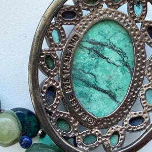 Peut inclure: Un pendentif ovale en argent sterling avec une grande pierre centrale verte veinée de noir, entourée de petites pierres bleues et vertes incrustées. Le cadre en métal martelé porte l'inscription BARSE 925 THAILAND.