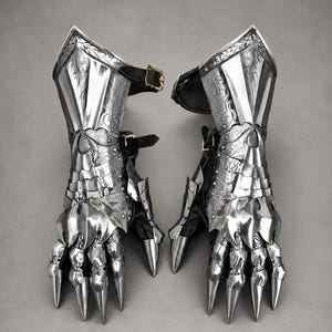 May include: A pair of polished silver metal gauntlets featuring intricate floral engravings on the forearm plates. The articulated finger sections end in sharp, pointed tips, and the pieces are secured with black leather straps and brass buckles.