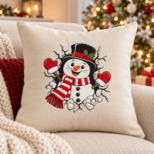 May include: A square beige throw pillow featuring an embroidered snowman wearing a black top hat with a red band and holly, a red and white striped scarf, and red mittens. The snowman appears to be breaking through a cracked wall surface.