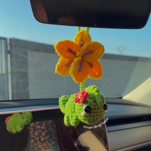 May include: A crochet car accessory featuring a bright yellow flower with purple center details hanging above a small green lizard figure. The lizard has a tiny pink flower on its head and black bead eyes. The text @rope.drop.loops is visible.