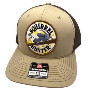 Puede incluir: Una gorra tipo trucker de color beige y marrón con un parche bordado circular en la parte delantera. El parche muestra una ardilla gris sobre una rama con hojas amarillas y el texto SQUIRREL HUNTER. Una pegatina negra en la visera dice RICHARDSON 112 TRUCKER.