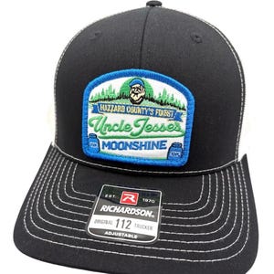 Puede incluir: Una gorra de camionero negra con costuras blancas y un parche rectangular con borde azul, un gráfico de bosque verde, un retrato de un hombre y el texto Hazzard County's Finest Uncle Jesse's Moonshine. Una pegatina Richardson 112 está en la visera.