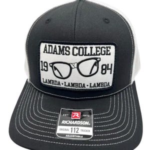 Può includere: Un cappellino trucker nero e bianco con una toppa rettangolare recante il testo ADAMS COLLEGE 1984 e un'illustrazione di occhiali. La toppa include il testo LAMBDA LAMBDA LAMBDA. Un adesivo Richardson 112 si trova sulla visiera nera.
