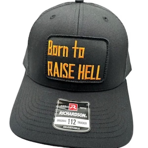 Puede incluir: Una gorra de camionero negra con un parche rectangular que presenta el texto bordado en naranja Born to RAISE HELL. La visera tiene una pegatina que dice RICHARDSON ORIGINAL 112 TRUCKER EST. 1970 ADJUSTABLE. La gorra tiene una parte trasera de malla.